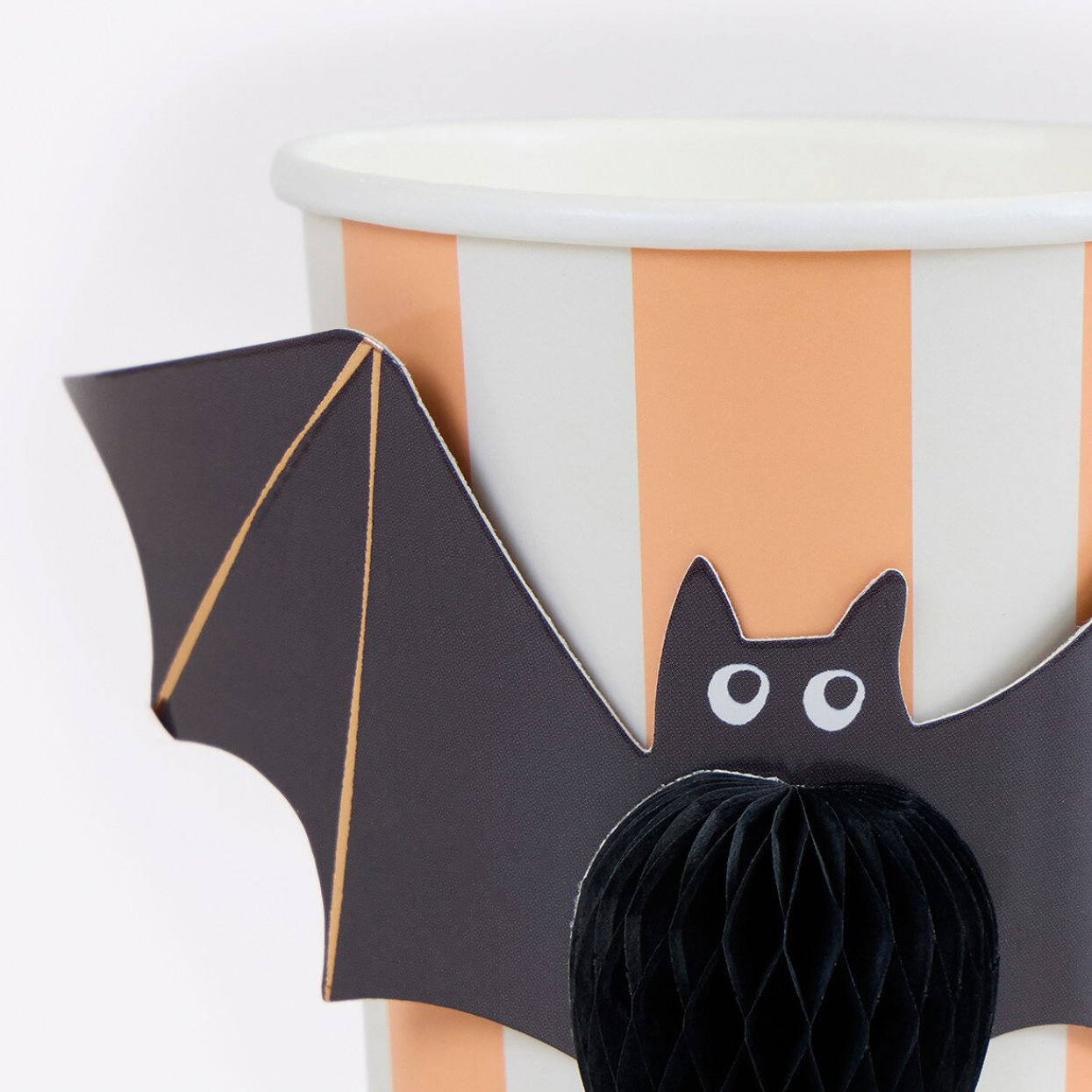 A close-up of a Halloween-themed party decoration featuring a black bat with white eyes and gold wing veins, attached to a white paper cup with peach and white vertical stripes, with a black honeycomb ball in front.