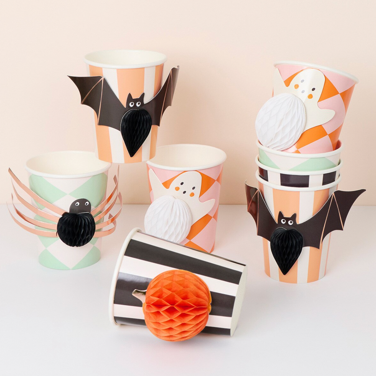 A set of Halloween-themed paper party cups and decorative spider and bat cutouts, arranged on a light beige background. The cups feature orange and white stripes, mint green and white patterns, black and white stripes, and illustrations of ghosts and spiders.