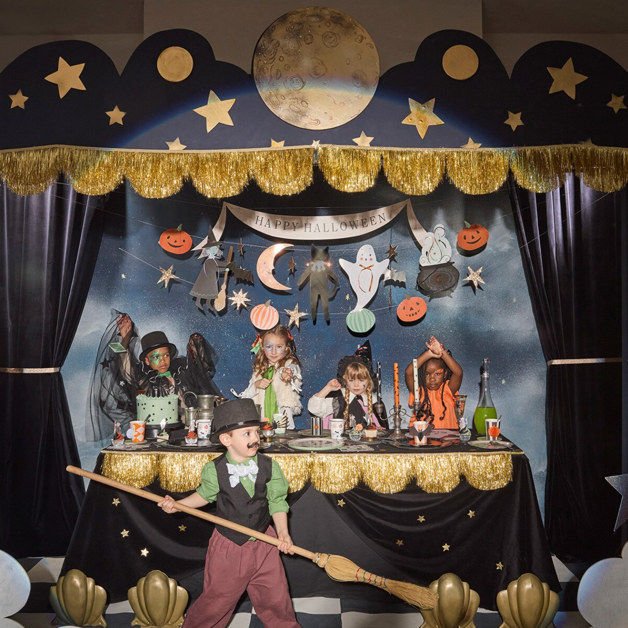 A Halloween-themed party backdrop setup with a dark blue starry sky background, gold fringe trim, black curtains, moon and star decorations, and a decorated table with spooky props. A child in costume stands in the foreground holding a broomstick.