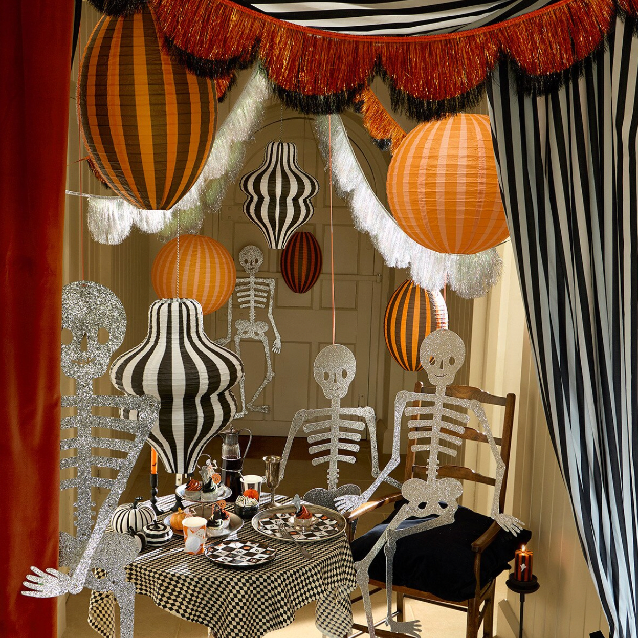 A Halloween-themed dining room scene decorated with skeleton cutouts, striped balloons, orange paper lanterns, fringe garland, and a checkered tablecloth.