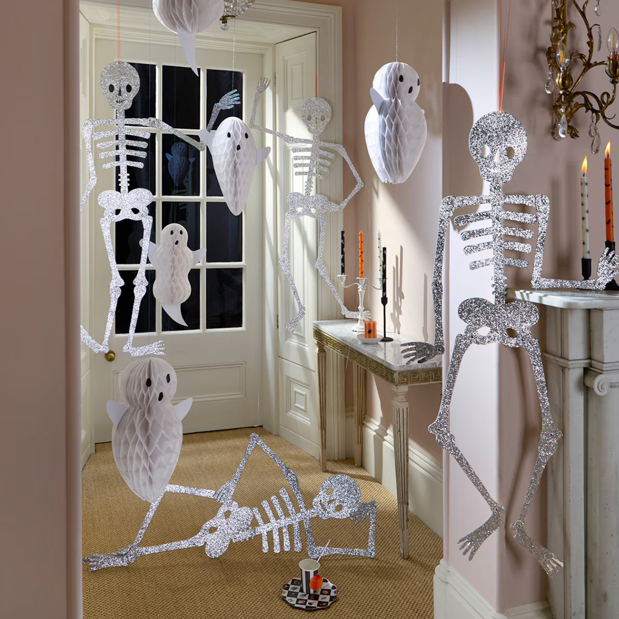 Halloween decorations featuring glittery silver skeleton figures and white paper-cut ghosts and owls hanging in a beige hallway with a fireplace and window.