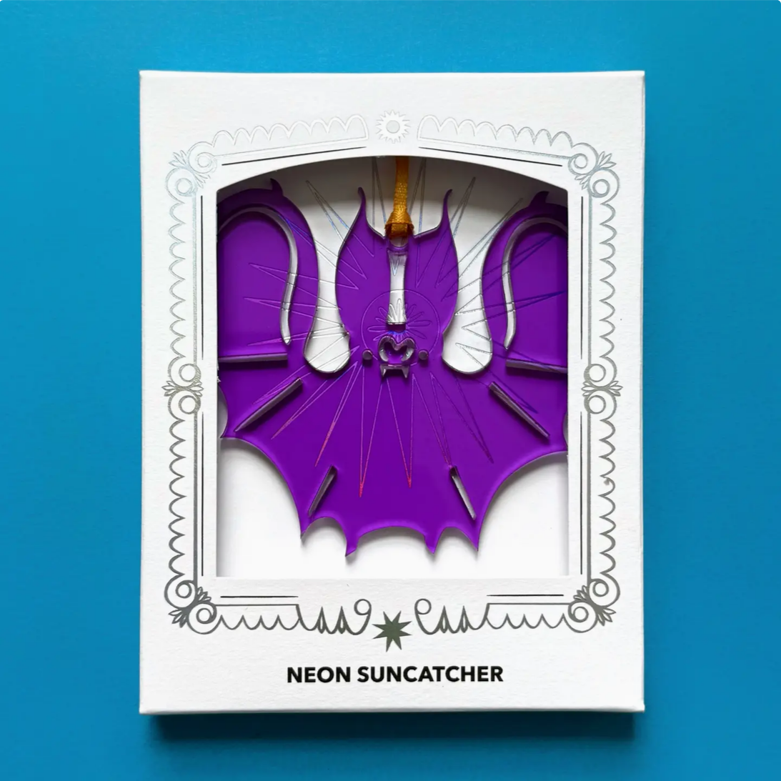 A neon purple bat-shaped suncatcher ornament displayed inside its white decorative window box packaging, set against a solid blue background.
