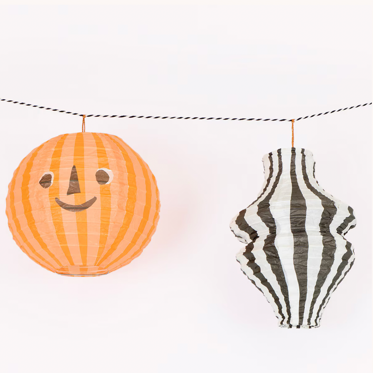 A Halloween hanging garland with two paper lanterns: one round orange lantern with a simple face and one black-and-white striped lantern, suspended by a string against a white background.