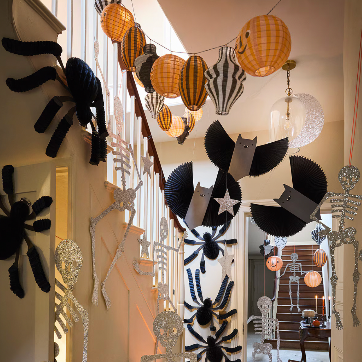 Halloween decorations hanging on a staircase landing, including black paper spiders, black-and-white striped paper lanterns, black paper bats, and silver skeleton cutouts.