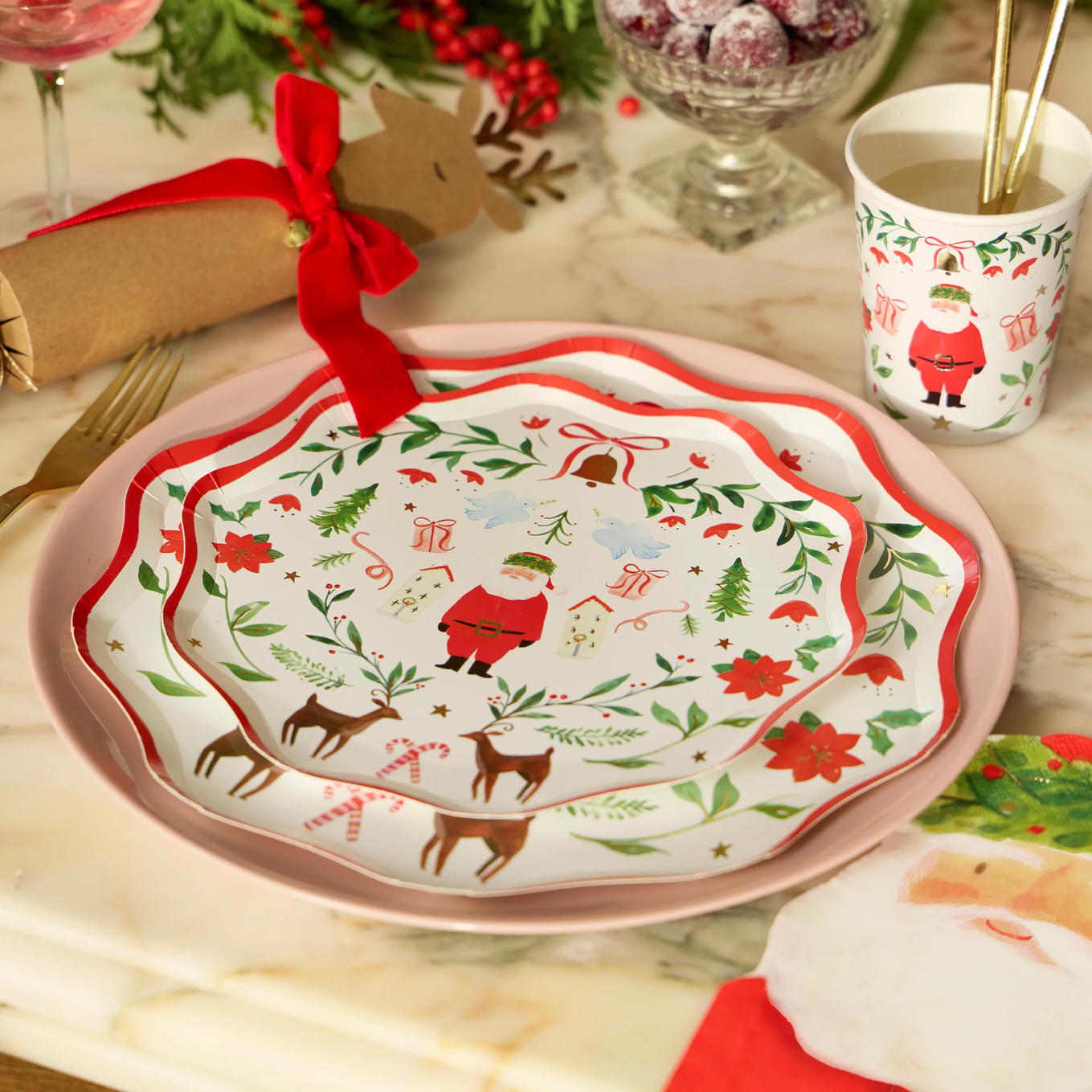 A set of Christmas-themed dinnerware, including a scalloped white plate with a pink rim and a matching cup, decorated with festive illustrations of Santa Claus, reindeer, pine trees, and red poinsettias on a marble tabletop.