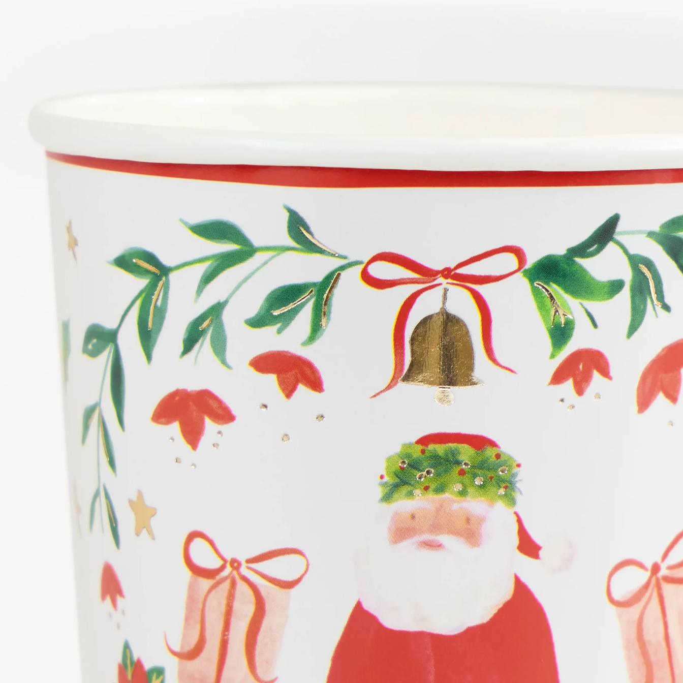 A close-up of a white ceramic Christmas cup featuring a festive print with a Santa Claus figure, green holly leaves, red bows, gold stars, and a gold bell.