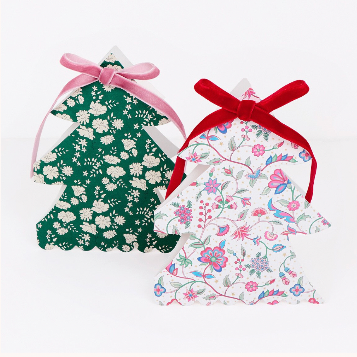 Two decorative Christmas tree-shaped paper ornaments or favors on a white background. The left one is dark green with a white floral print and a pink ribbon bow.