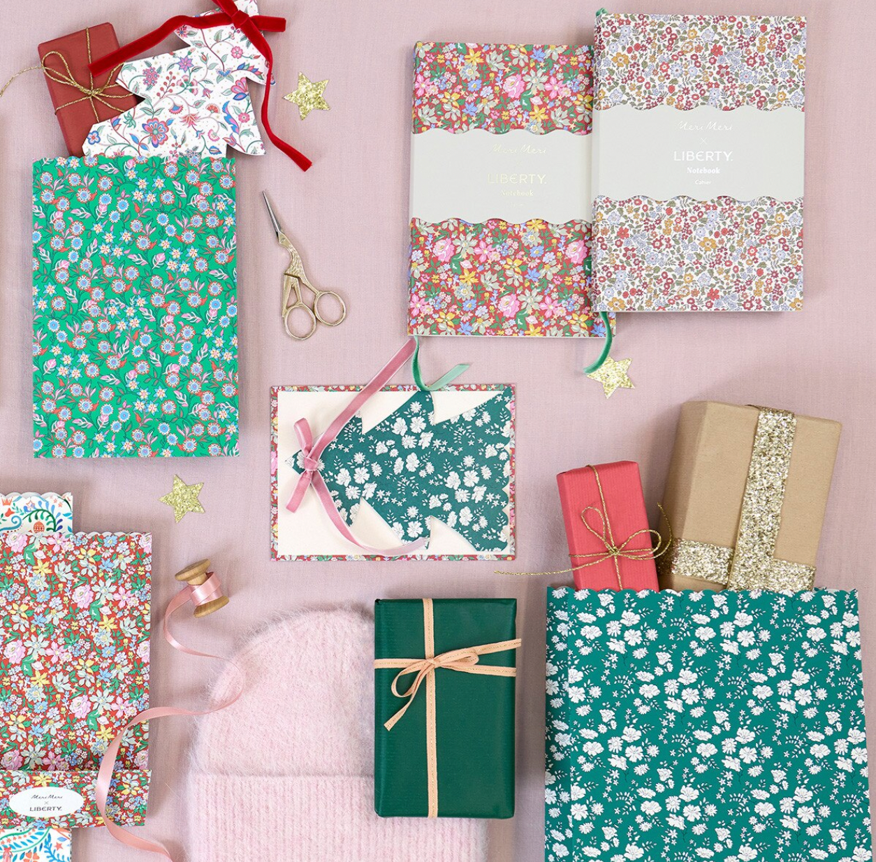 A styled flat-lay of gift-wrapping supplies and wrapped presents on a pale pink background, including floral and speckled wrapping paper, ribbon, scissors, tape, and several gift boxes.