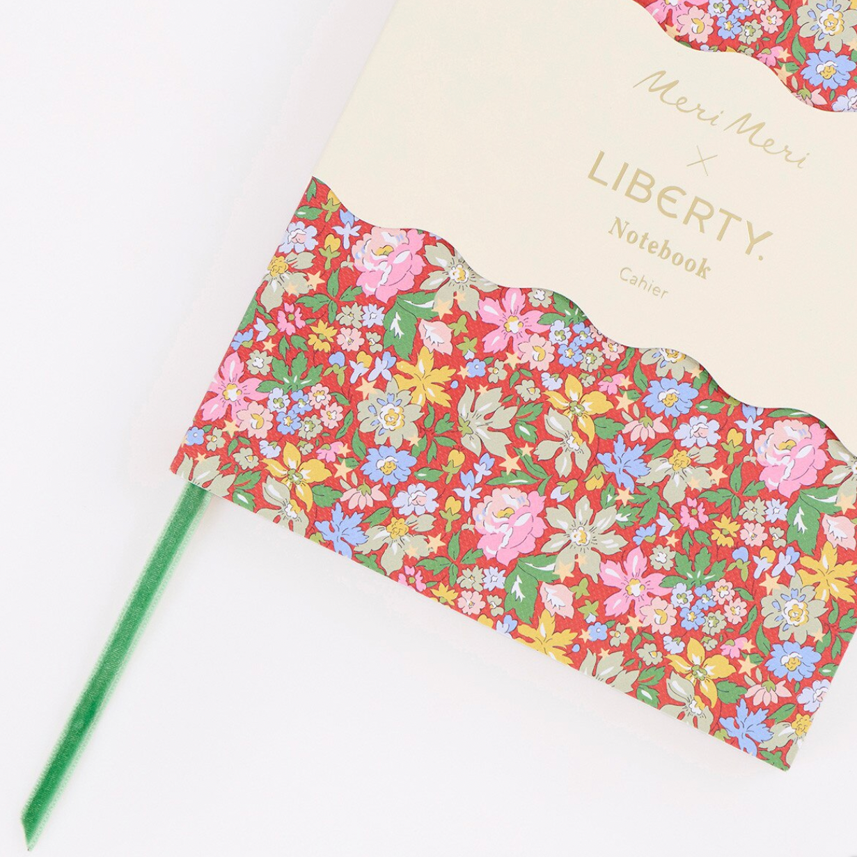 A notebook with a cream-colored top section and a scalloped floral patterned lower section, shown on a white background with a green pen resting beside it. The cover includes the text 'Menu Meri x LIBERTY Notebook'.