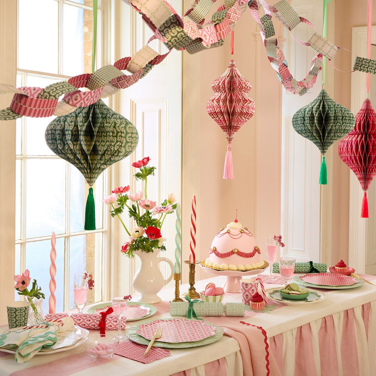 A styled party table decorated with pink and green patterned paper lanterns hanging from a garland strung across a windowed wall. The table is set with white tablecloths, pink and white plates, striped candles, and a pink tiered cake stand.