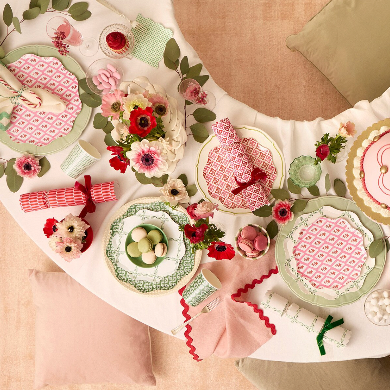 A beautifully styled table setting featuring a white tablecloth with a green and pink floral vine pattern, coordinated plates, pink napkins with red patterned borders, red and white striped crackers, and floral centerpieces.