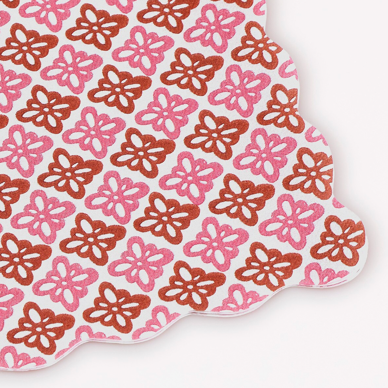 A scalloped-edged paper napkin featuring a repeating  floral pattern in pink and brown on a white background.