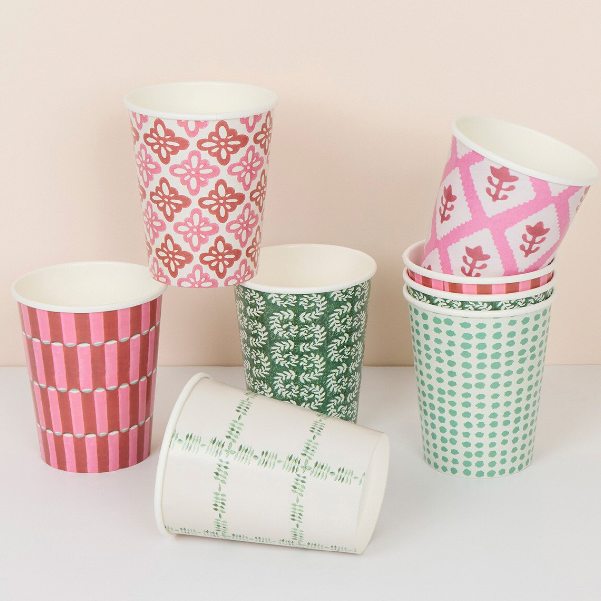 A set of six reusable paper cups with various colorful patterns, including pink and white stripes, green and white floral motifs, pink and white geometric designs, and green polka dots, arranged on a light surface against a pale beige background.