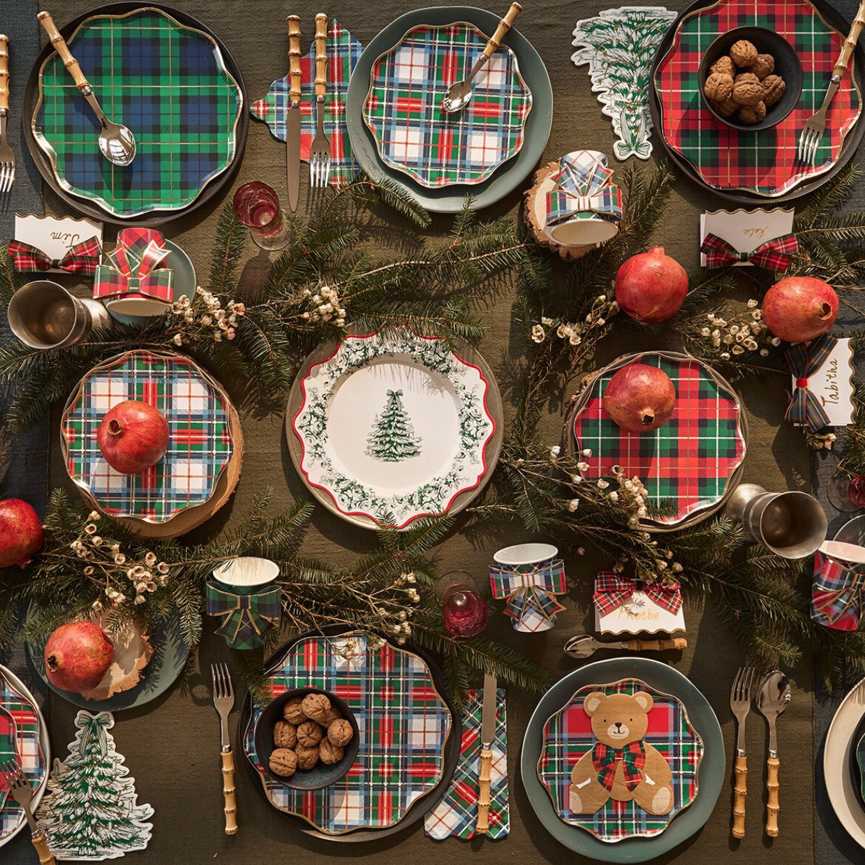 A styled holiday table setting featuring scalloped plaid plates in red, green, and blue tartan patterns, matching napkins, wooden-handled cutlery, pine garlands, red apples, and small decorative ornaments on a dark tabletop.