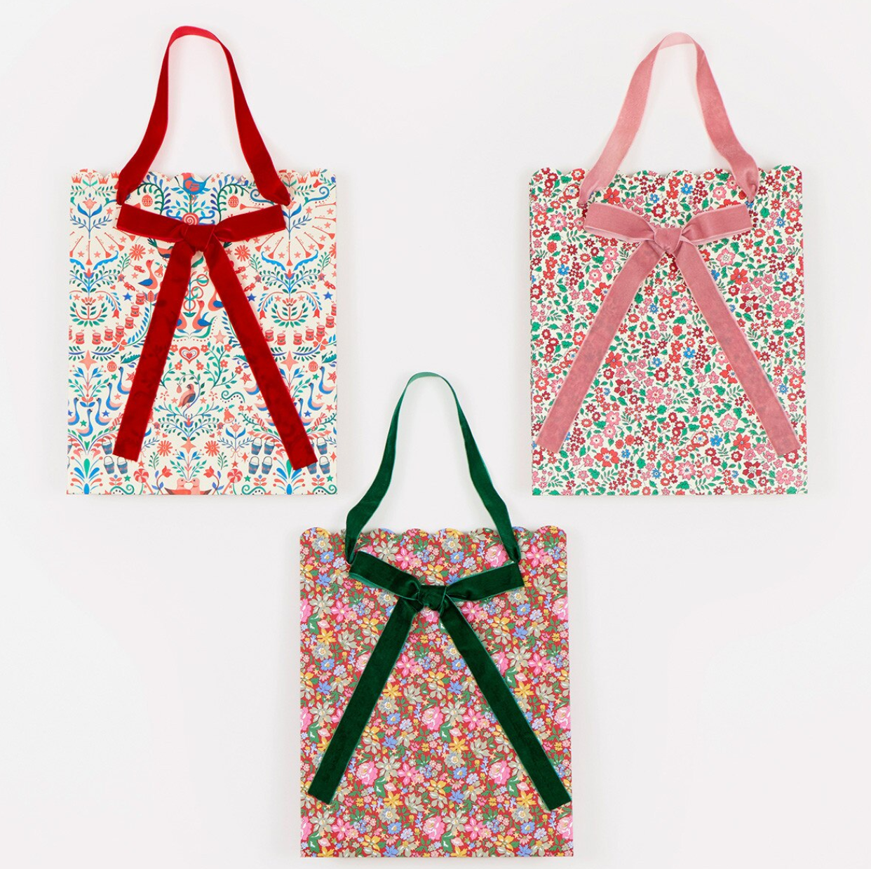 Three fabric tote bags with floral prints and ribbon handles, arranged on a white background. The top-left bag has a red ribbon handle, the top-right has a pink ribbon handle, and the bottom-center has a dark green ribbon handle.