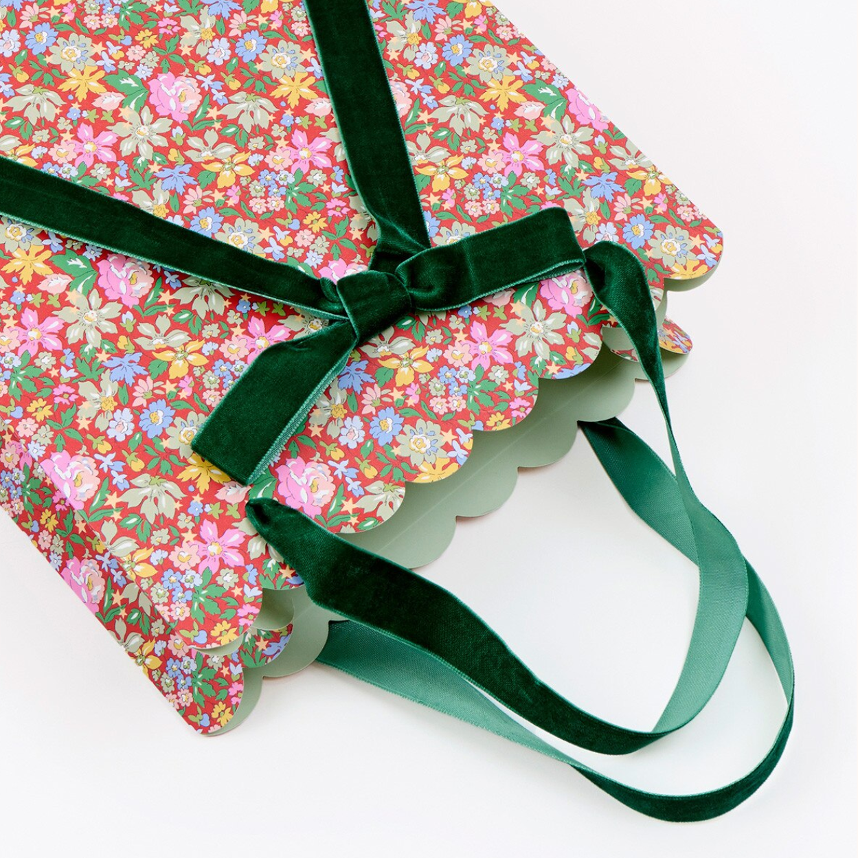 A gift bag with a pink and multicolor floral print, scalloped edges, and dark green velvet ribbon handles tied in a bow, shown on a plain white background.