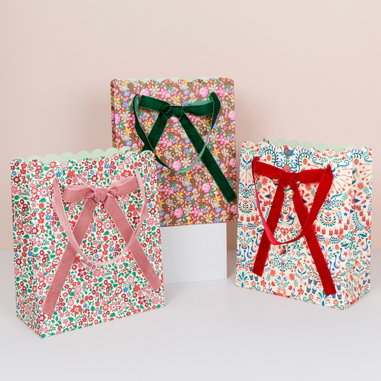 Three floral gift bags with ribbon handles arranged on a white surface against a pale beige background. The left bag has a pink ribbon, the center bag has a dark green ribbon, and the right bag has a red ribbon.