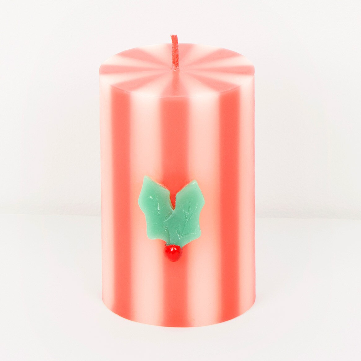 A pink striped pillar candle with a green holly leaf and red berry ornament on the front, set against a plain white background.