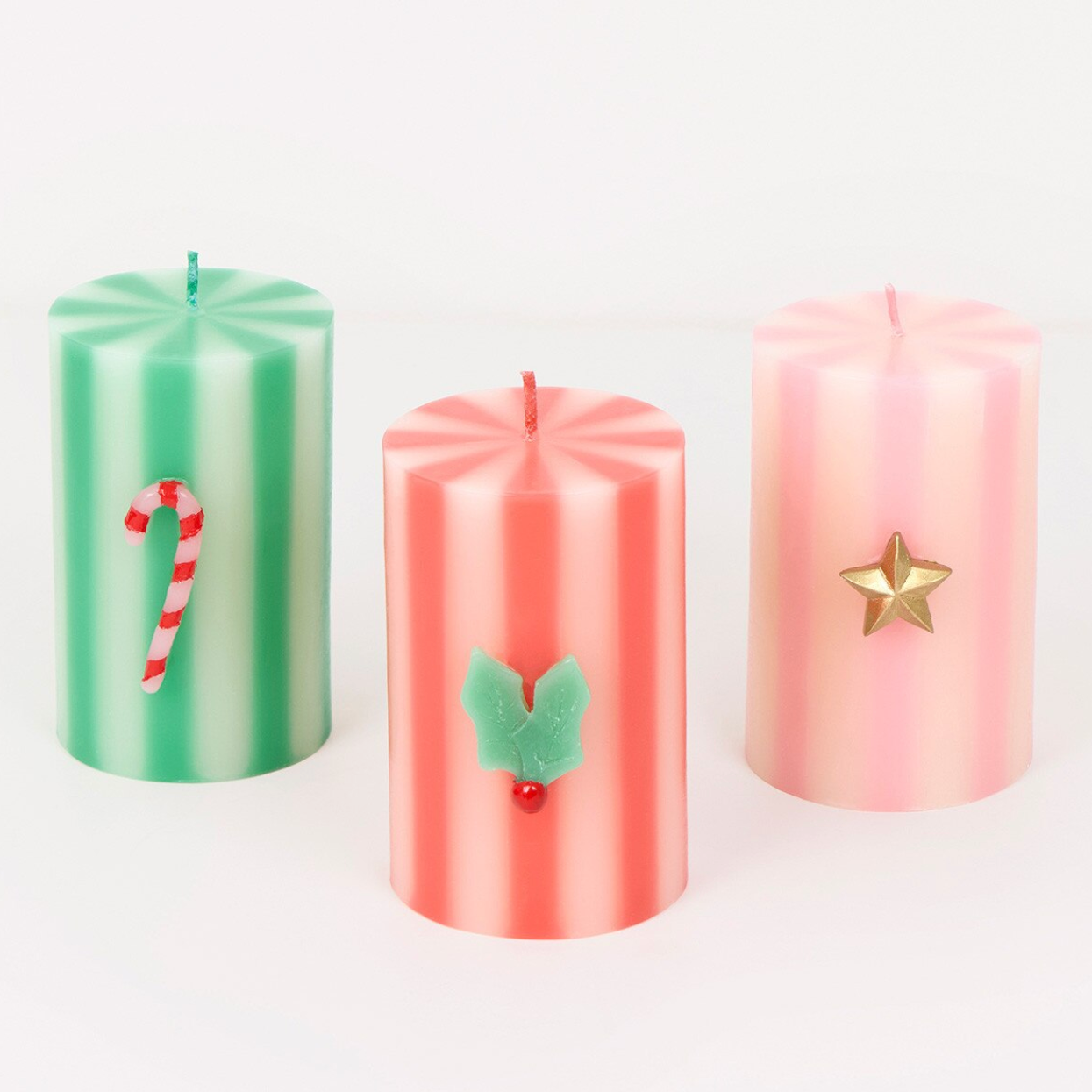 Three striped Christmas pillar candles on a white background: one mint green with a candy cane decoration, one coral pink with holly leaves and a red berry, and one light pink with a gold star.