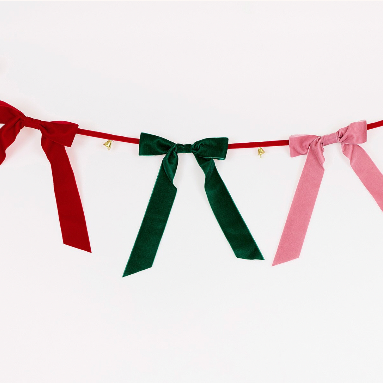 A festive velvet ribbon bunting garland with three bows in red, dark green, and light pink, strung along a thin red cord with small gold bells.
