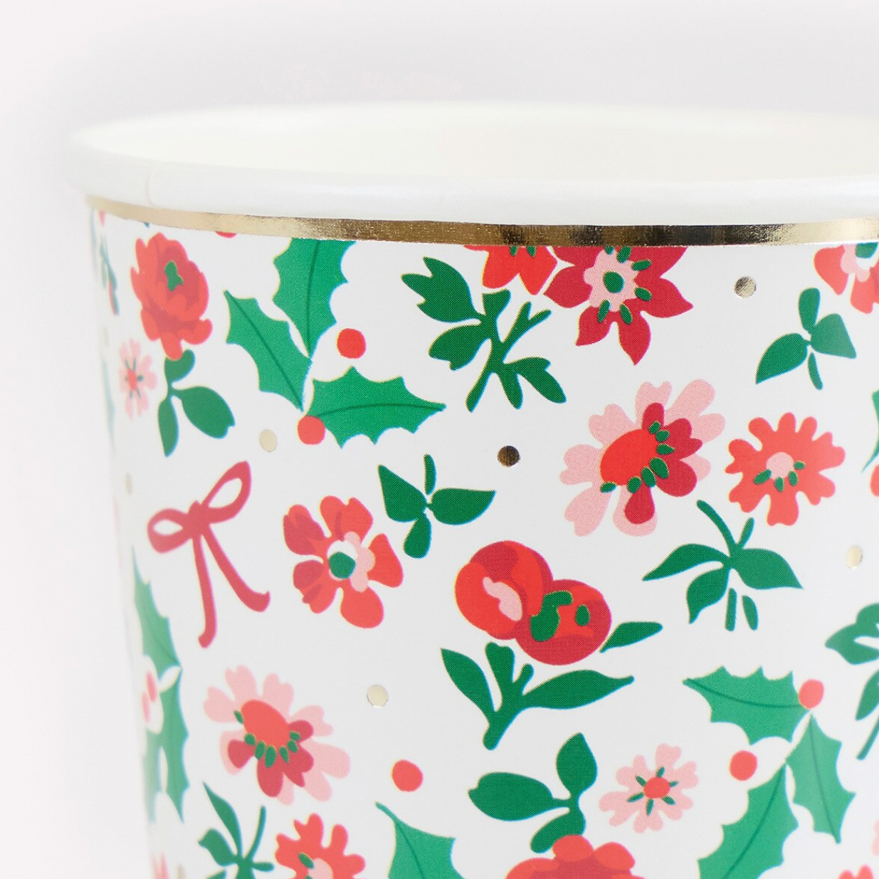 A close-up of a white cylindrical container or cup with a festive Christmas print featuring red and pink flowers, green holly leaves, and gold polka dots, finished with a metallic gold band near the rim.