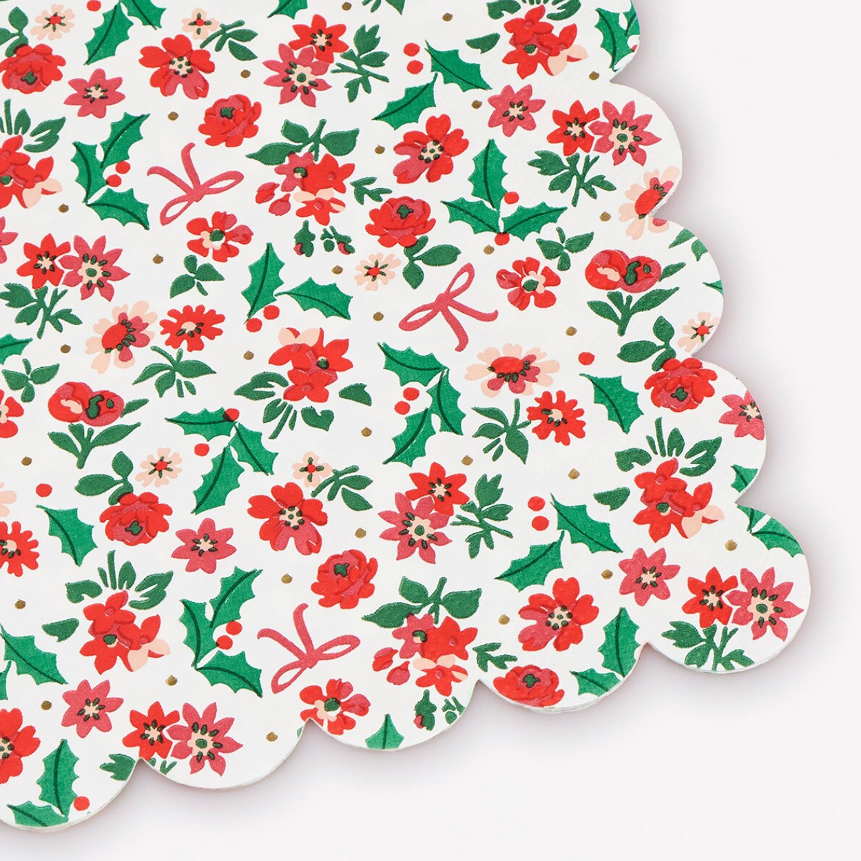 A scalloped-edge paper napkin or table linen featuring a festive Christmas floral print on a white background, with red poinsettias, green holly leaves, small red bows, and tiny gold dots.