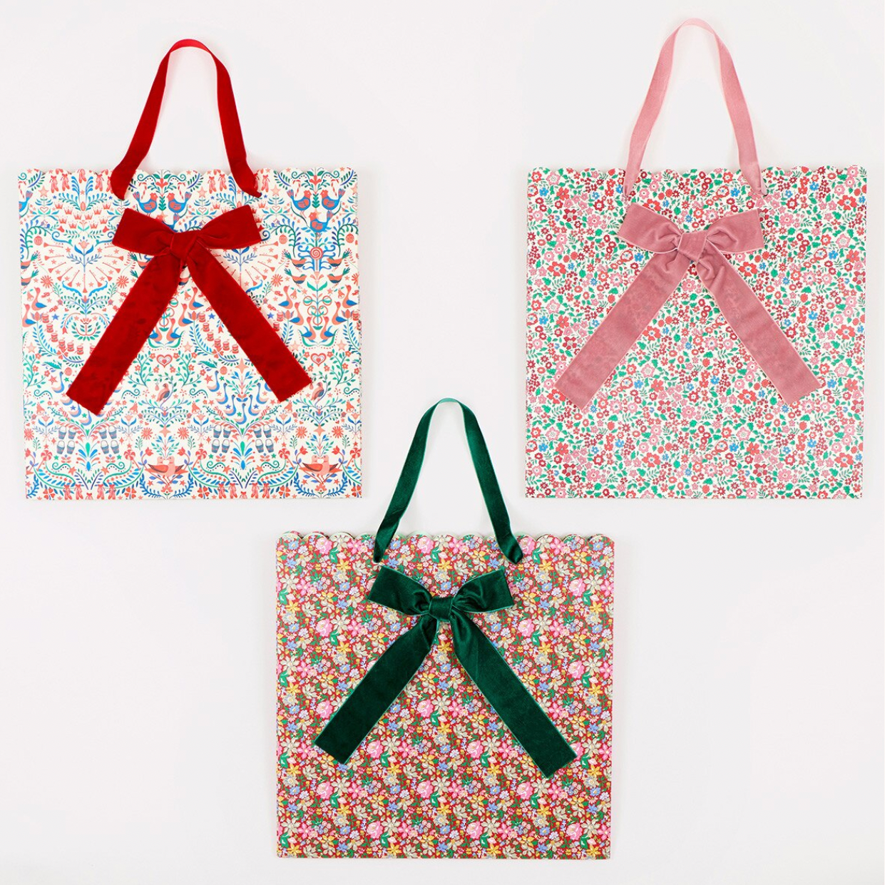Three fabric tote bags with floral prints and velvet ribbon handles, arranged on a white background. The top-left bag has a red ribbon handle, the top-right has a pink ribbon handle, and the bottom-center has a dark green ribbon handle.