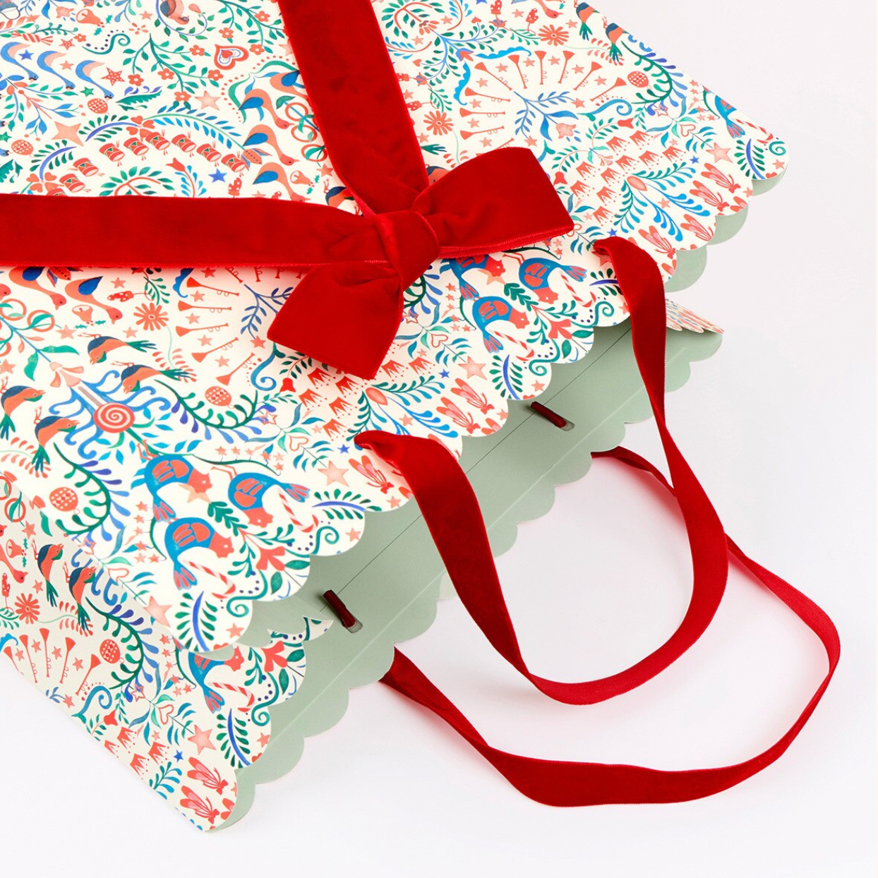 A gift bag with a colorful folk-art patterned body, scalloped edges, and long red velvet handles tied with a red bow, shown on a white background.
