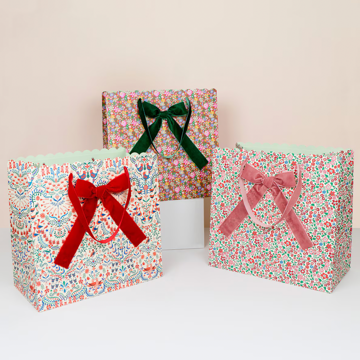 Three floral gift bags and matching gift boxes with ribbon handles, displayed on a white surface against a light beige background. The left bag has a red ribbon, the right bag has a pink ribbon, and the back box has a dark green ribbon.