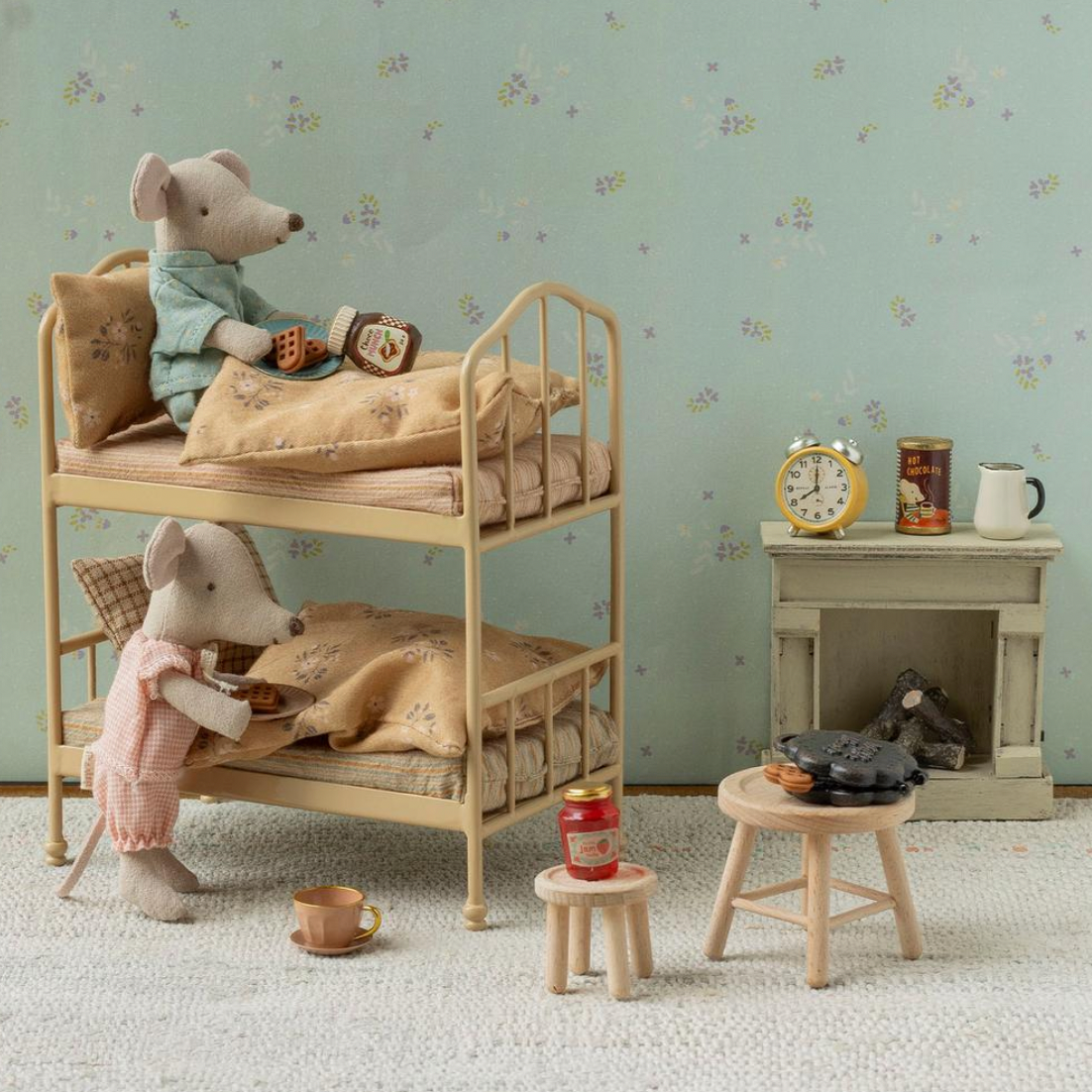A styled miniature dollhouse interior scene with two plush mouse figures arranged on a wooden bunk bed, alongside a small table, stool, mug, and fireplace display against floral wallpaper.