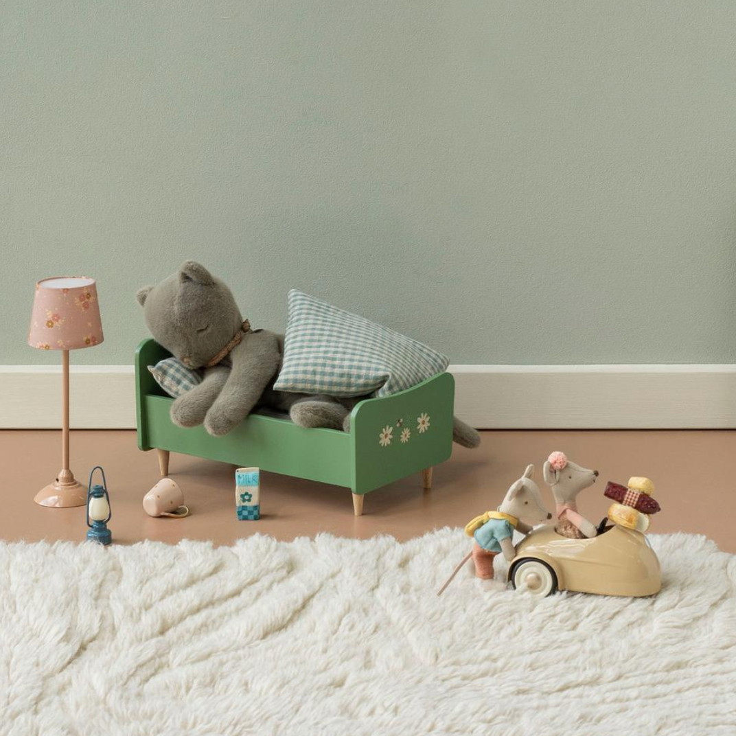 A styled children's room scene with a green wooden bed, plush gray teddy bear, checkered pillow, small pink lamp, green cabinet, and a small toy vehicle on a white shag rug against a pale green wall.