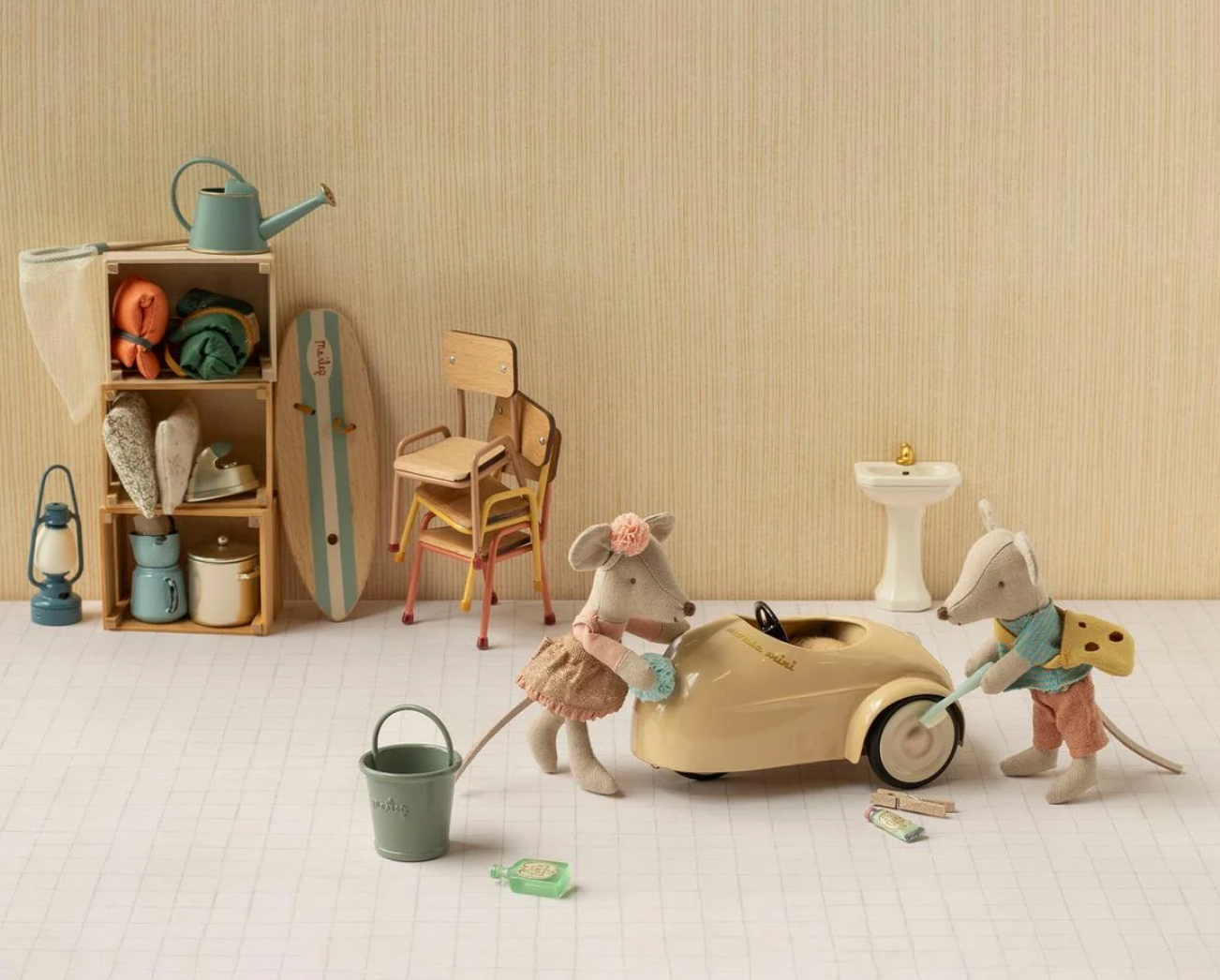 A styled miniature dollhouse or toy room scene with animal characters around a small beige car, shelves with toys, a chair, lanterns, and bathroom accessories, set against a wood-grain wall and white floor.