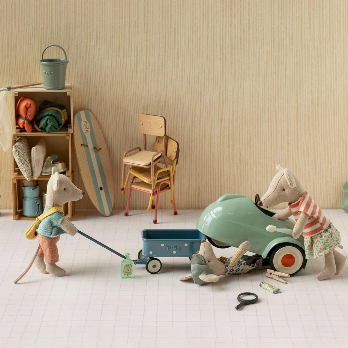 A styled toy scene featuring plush animal figures and a vintage toy car in a room with beige wallpaper, shelves, a surfboard, and a small blue wagon.