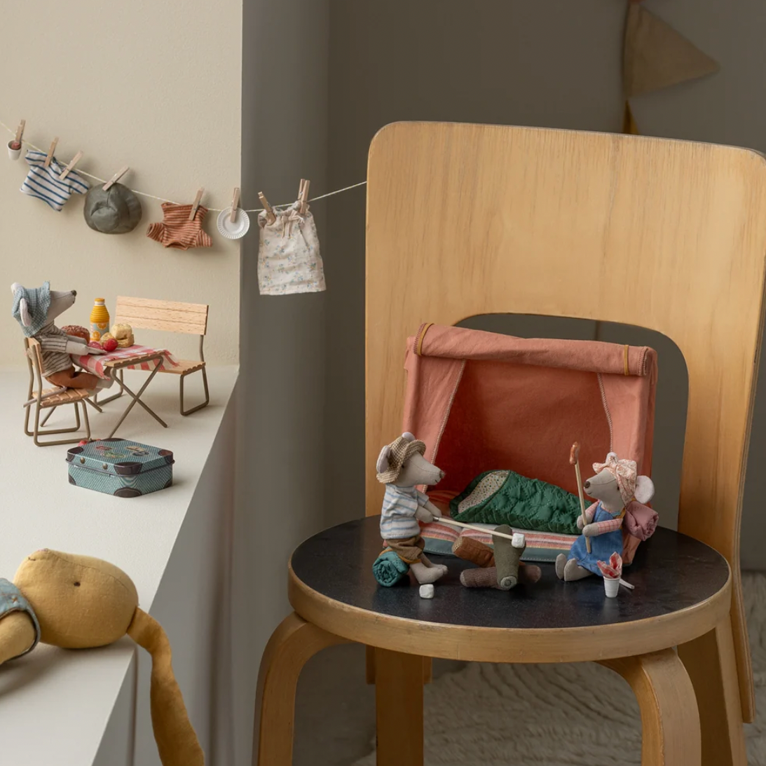 A styled interior scene showing a small wooden chair with a round dark seat, holding a miniature dollhouse campsite setup with tiny animal figures and a pink tent. The room has warm neutral walls, a hanging clothesline with small accessories, and other miniature decor in the background.