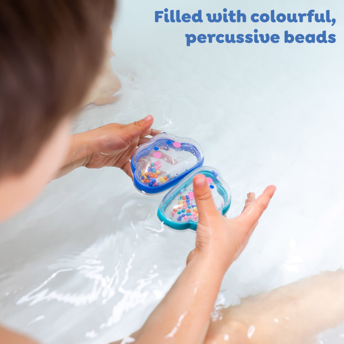 A person holding two heart-shaped blue plastic bath toys filled with colorful percussive beads in a white bathtub, with the text 'Filled with colourful, percussive beads' in blue at the top right.