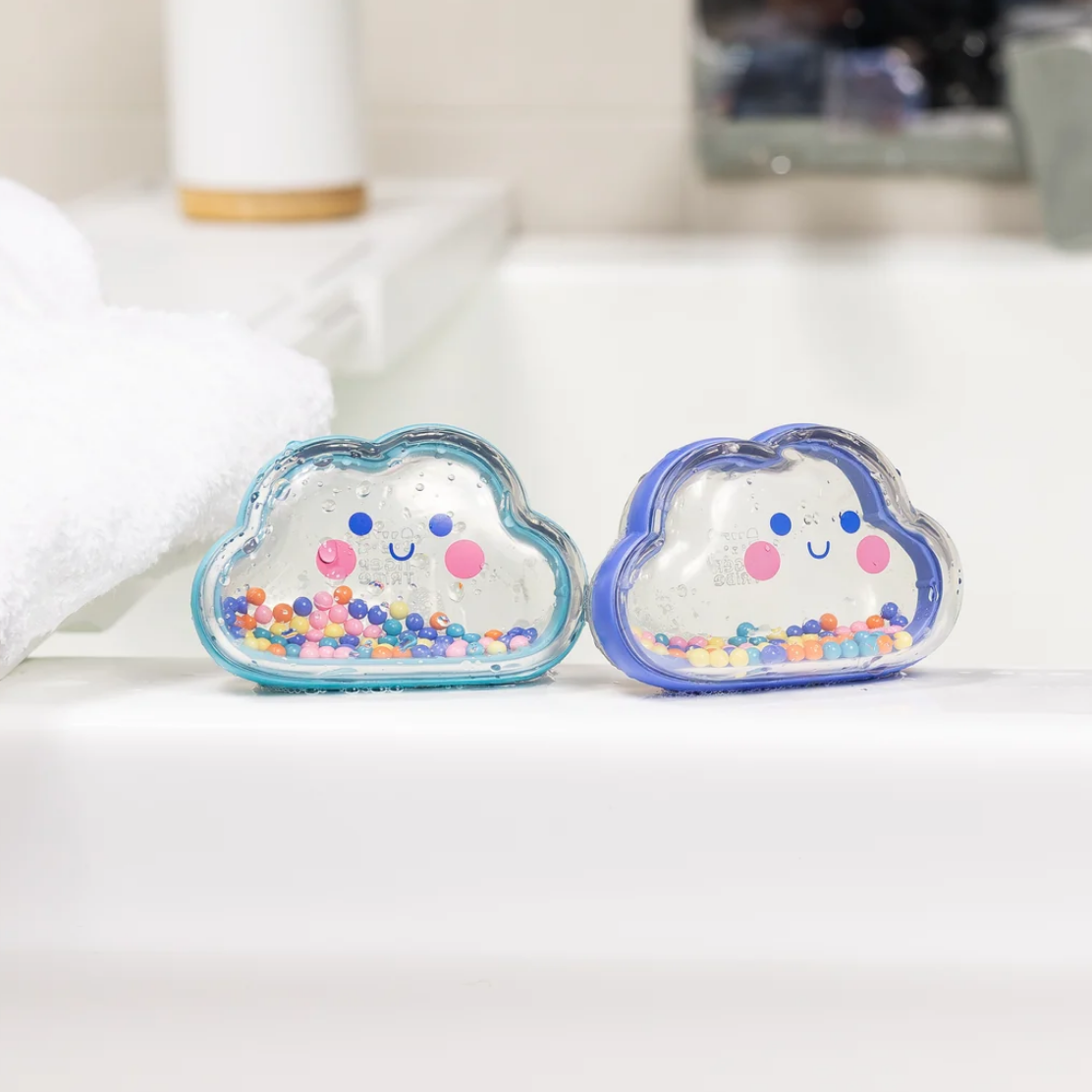 Two cloud-shaped bath bombs with smiling faces and colorful beads inside, resting on a white bathroom counter next to a folded white towel.