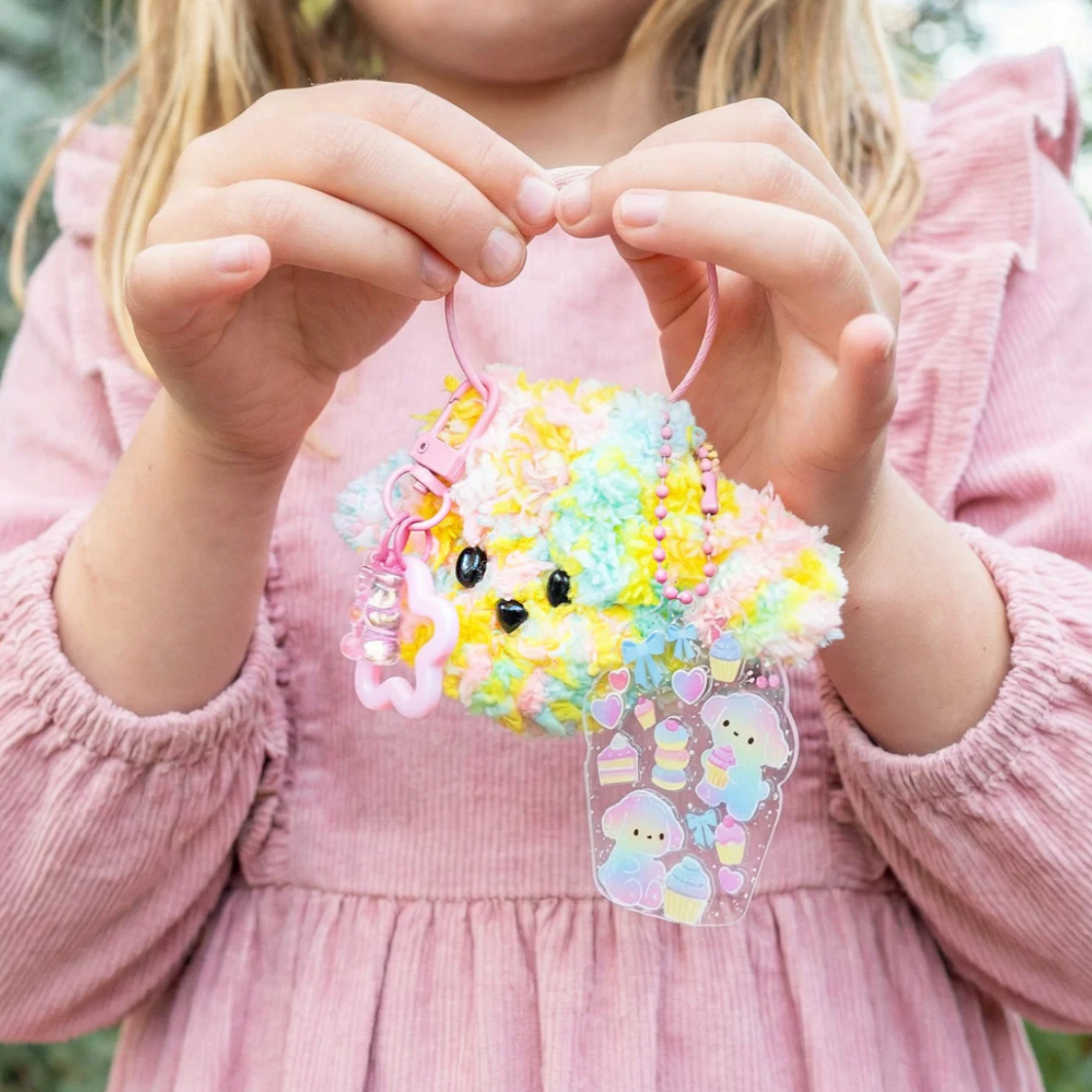 A child in a pink corduroy dress holds a pastel multicolor fluffy keychain charm with a clear sticker tag featuring cute characters.