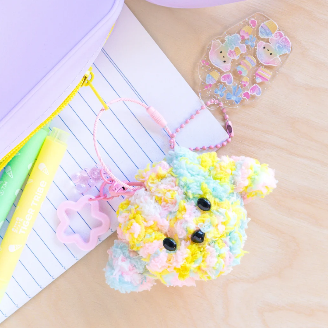 A pastel multicolor fluffy pom-pom character keychain or phone bag charm with black bead eyes, attached to a pink beaded strap and small pink flower charm, styled on a notebook with stationery props.