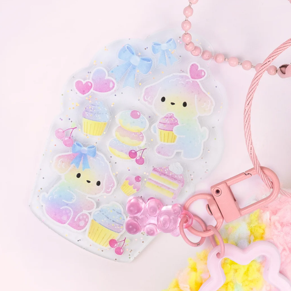 A pastel pink and iridescent acrylic keychain bag decorated with cute cartoon characters, cupcakes, and hearts, shown with a pink bead chain and clasp on a light pink background.
