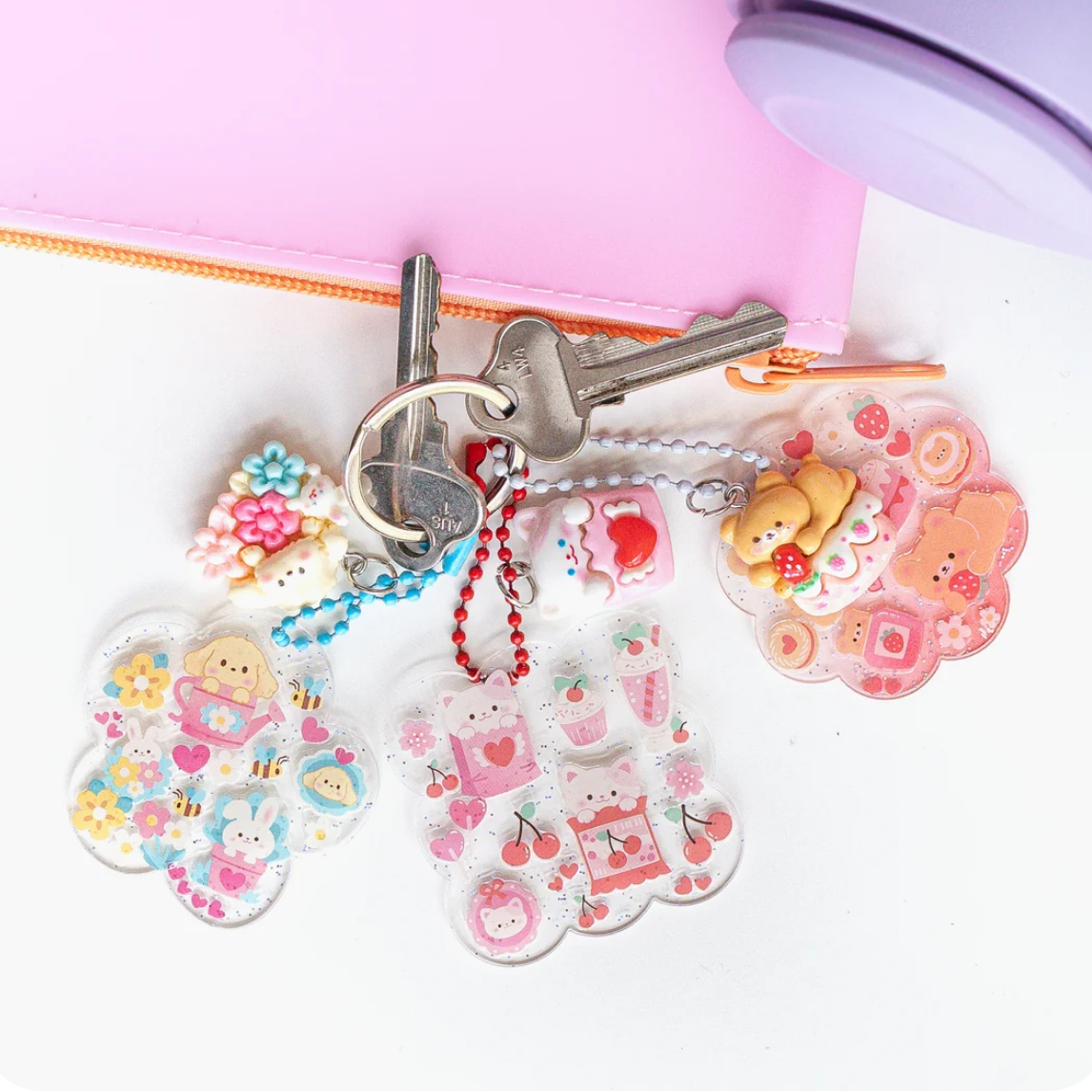 Three pastel pink and multicolor acrylic keychains with cute cartoon characters, shown attached to metal keys on a white surface next to a pink zip pouch.