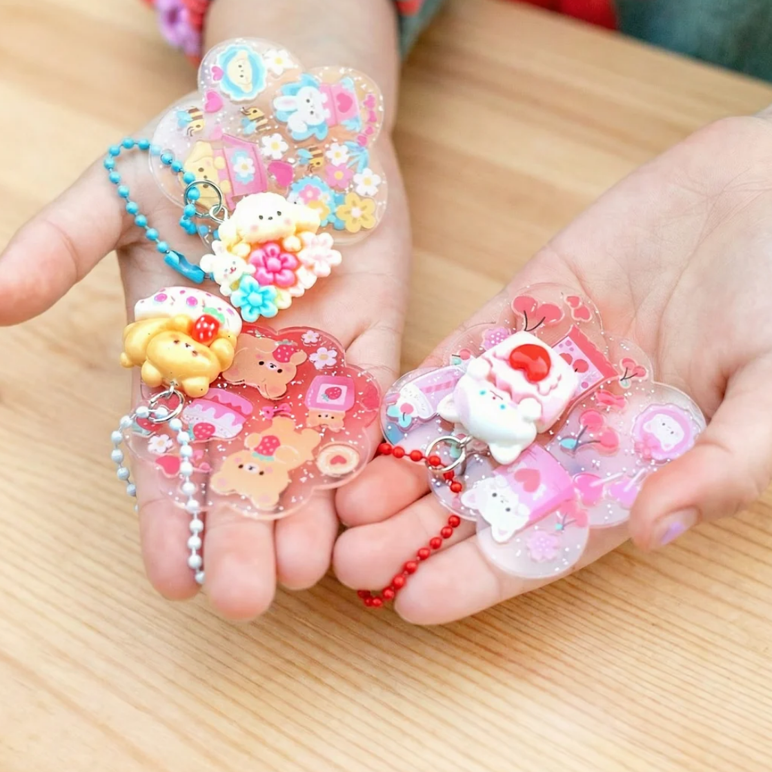 Two hands holding several small clear acrylic charms decorated with pastel cartoon characters, flowers, hearts, and bead keychains, set against a light wood tabletop.