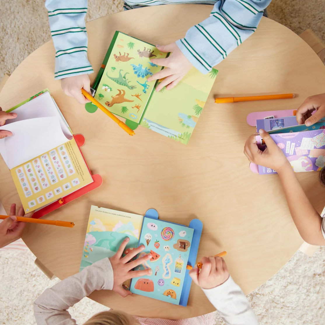 Overhead view of children's hands interacting with colorful educational activity books and orange markers on a round light wood table.