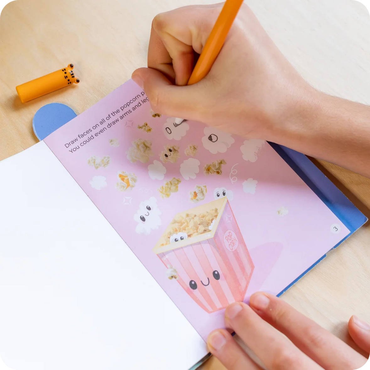 A child's hand holding an orange pencil, drawing on a pink children's activity book with cute cartoon popcorn illustrations. The book is open on a light wooden desk, with a small yellow and blue toy accessory visible in the upper left.
