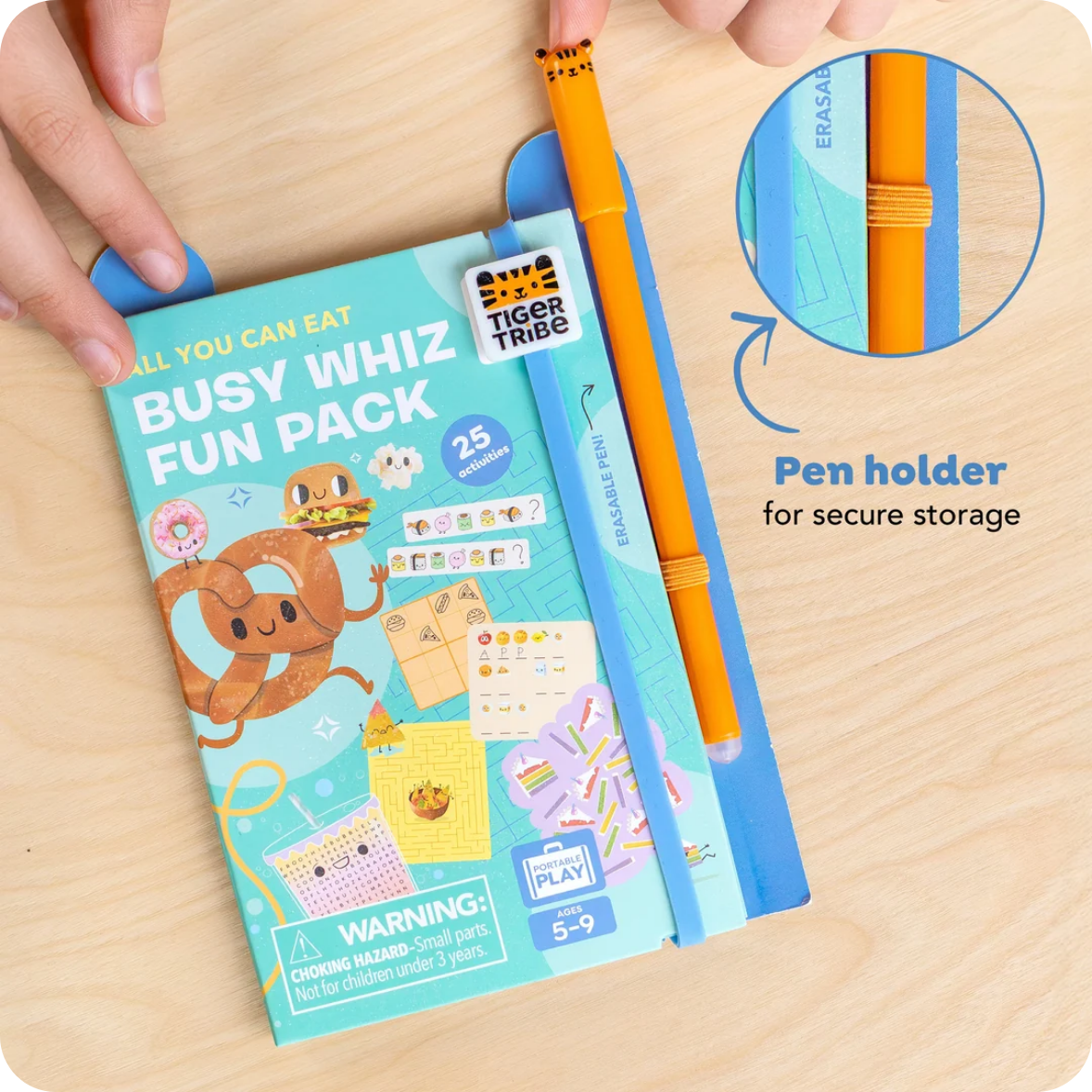 A hand holds a colorful 'Busy Whiz Fun Pack' notebook on a light wood desk while an orange pen rests inside a blue pen holder attached to the pack. A circular inset shows the pen holder in detail, and text reads 'Pen holder for secure storage'.
