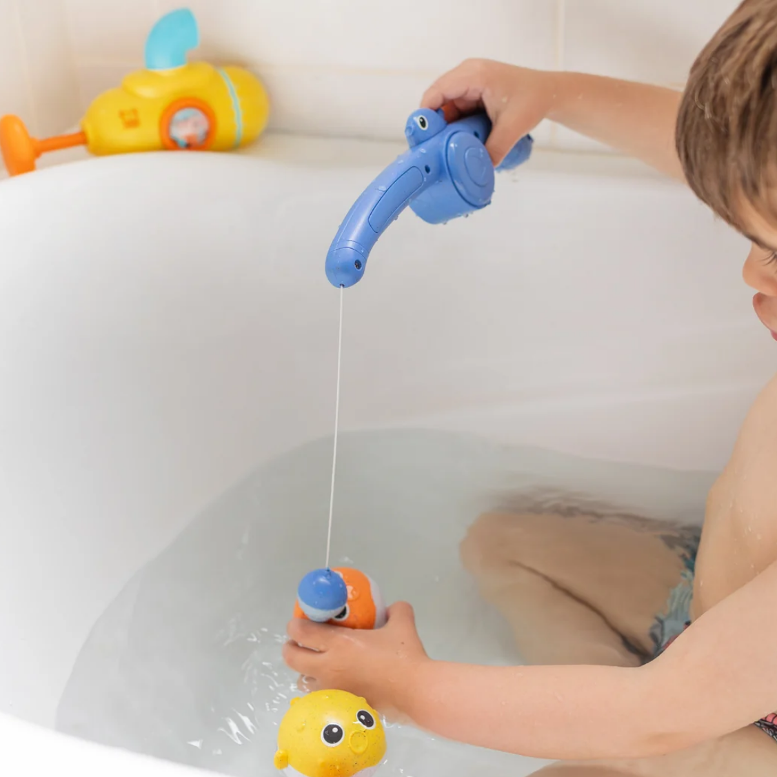 A child sitting in a bathtub uses a blue toy fishing rod with a string to try and catch a yellow rubber duck. A yellow and blue toy submarine is visible in the tub on the left.
