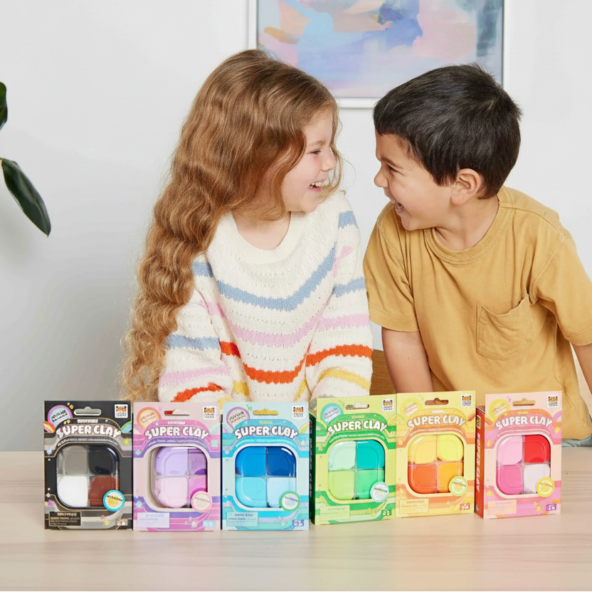 A child's play set featuring five colorful Super Clay air-dry modeling kits lined up on a table, with a young girl and boy smiling behind them against a light wall with a colorful painting.