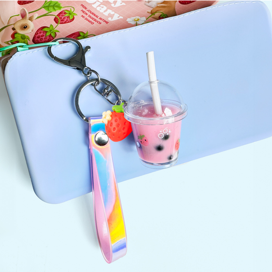 A pastel iridescent wrist strap keychain with a black clasp, attached to a metal key ring. The keychain includes a small pink boba tea cup charm with a clear dome lid, white straw, and a red strawberry accent, displayed against a light blue notebook.