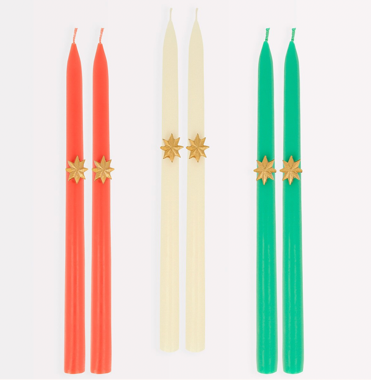 Four taper candles arranged in three pairs on a white background: two coral-red candles, two cream candles, and two green candles. Each pair has a small gold starburst detail near the top.