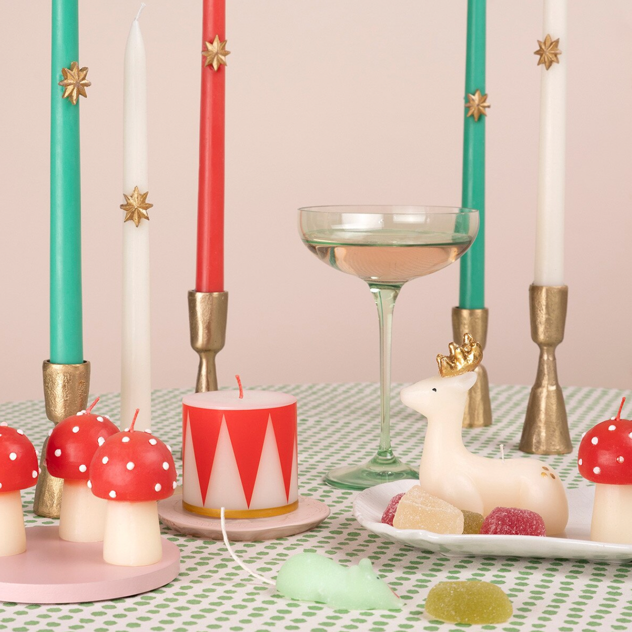 A styled tabletop scene with decorative candles, a martini glass, a white deer figurine, mushrooms, candies, and a small candle on a green-and-white checkered cloth against a pale pink background.