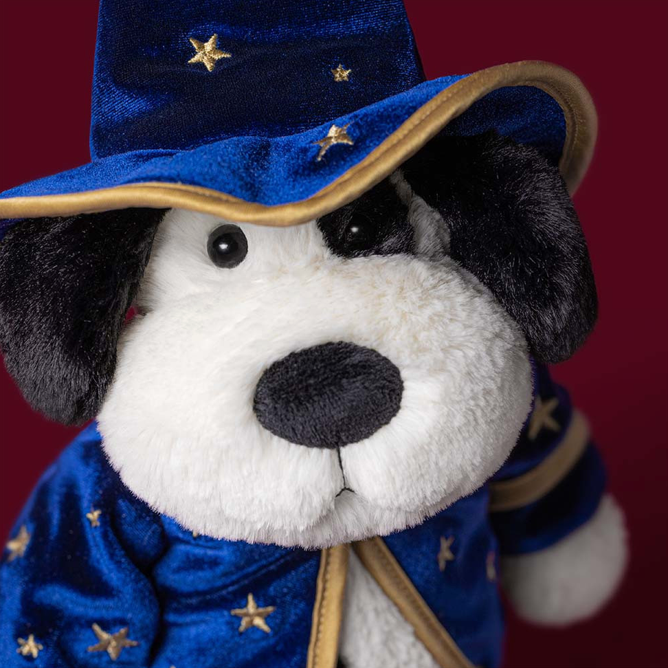A plush dog toy dressed as a wizard, featuring a white and black face, a blue velvet-like hat with gold star details, and a matching blue robe with gold trim, set against a dark red background.